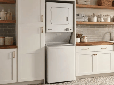 A stacked white washer and dryer set between white cabinets and wooden shelves